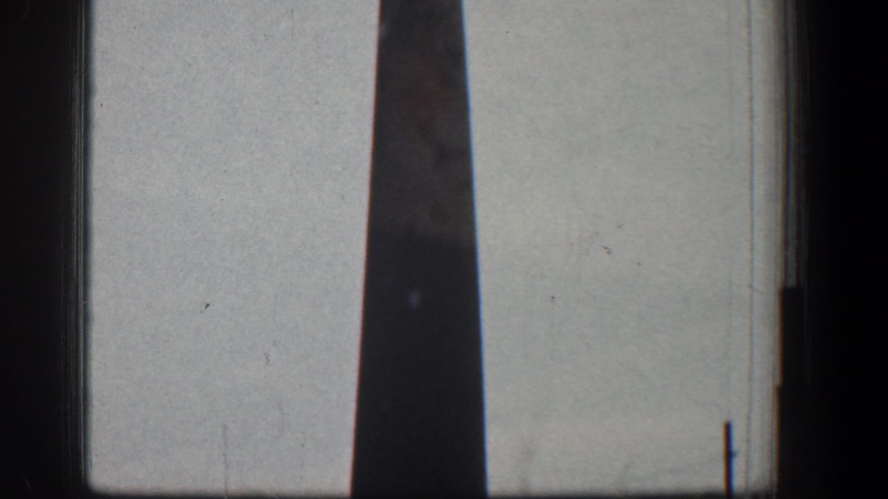 Narrow dark chimney or stack seen through moving-vehicle framing against a pale sky.