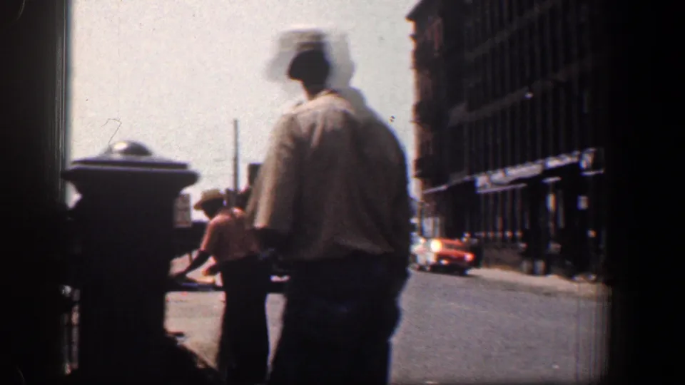 1961 Baltimore sidewalk view with a man in a hat walking near pedestrians, cars, a signpost, and tall urban buildings.
