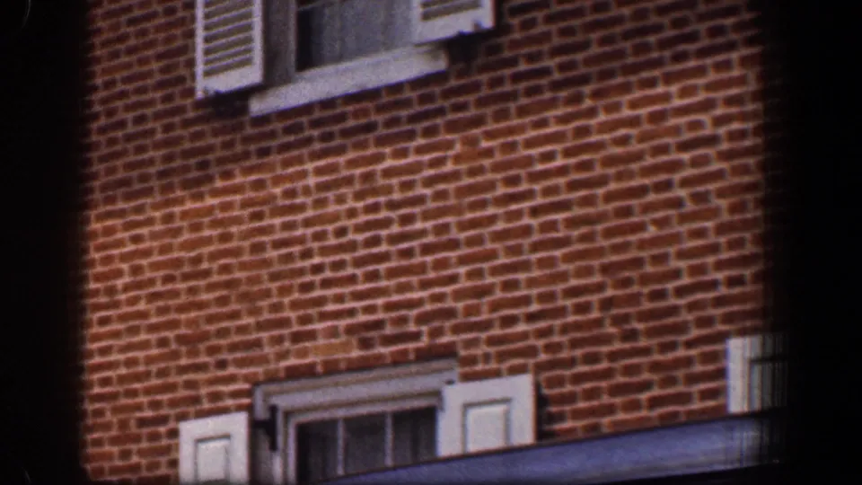 1961 Baltimore frame showing a brick building facade, shuttered windows, and the edge of a passing dark vehicle or obstruction.