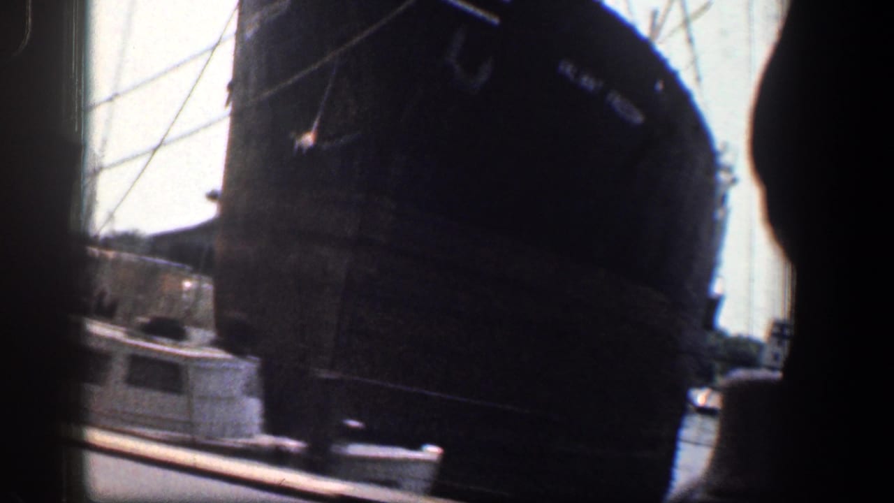 Large dark ship beside a waterfront street with another vessel and film gate edges visible.