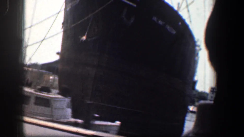 1961 Baltimore waterfront view from a moving vehicle, with a large dark ship, another harbor vessel, street edge, and bright sky.