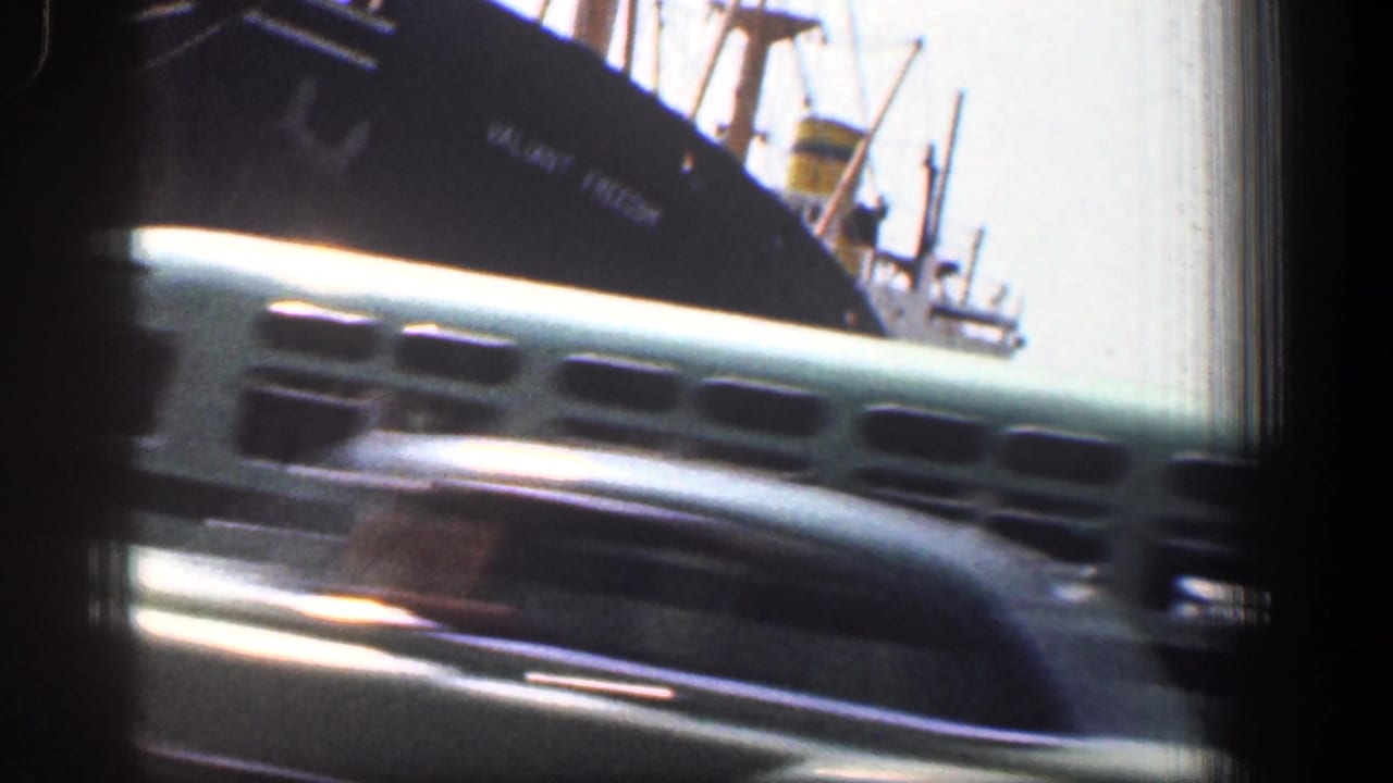 Light green bus passing in front of a large dark ship at the waterfront.