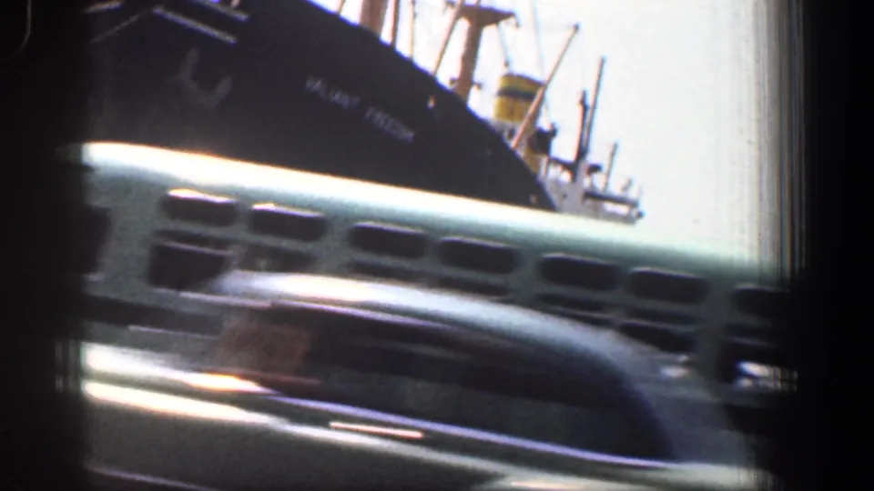 A light green bus moves across the foreground while a dark harbor ship and masts remain behind it in 1961 Baltimore.