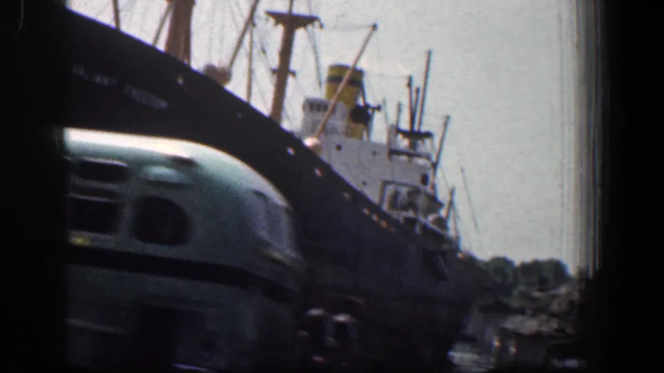 The moving camera catches the front of a light green bus near docked vessels, rigging, and waterfront structures in Baltimore.