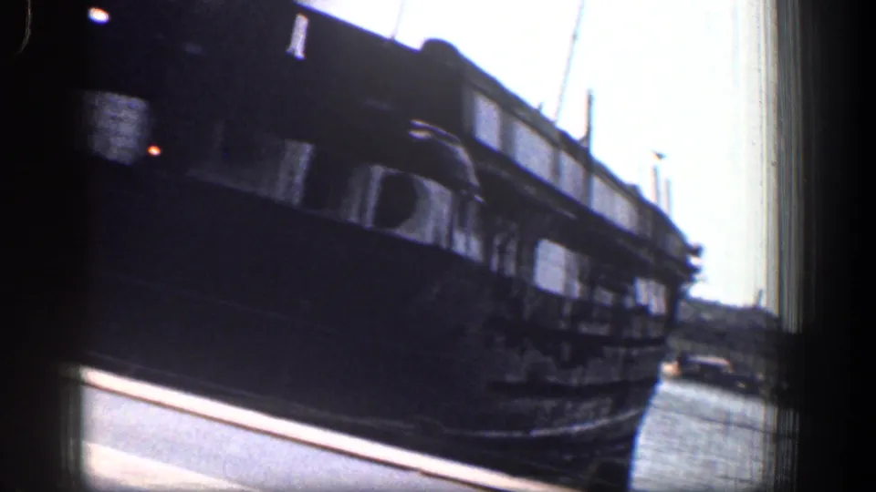 A dark ship hull fills the right side of the frame as the camera looks along a waterfront road with distant cars and harbor structures.
