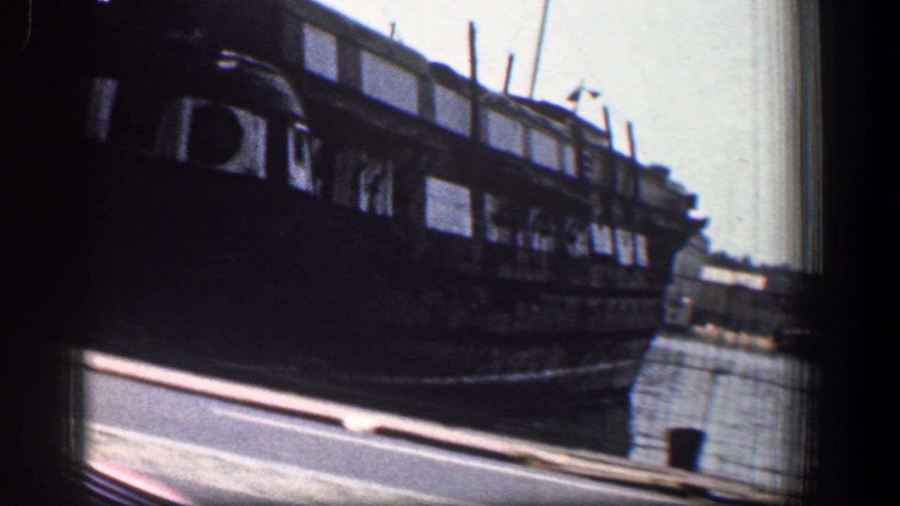 Docked vessel beside harbor water, filmed from a moving roadside viewpoint.