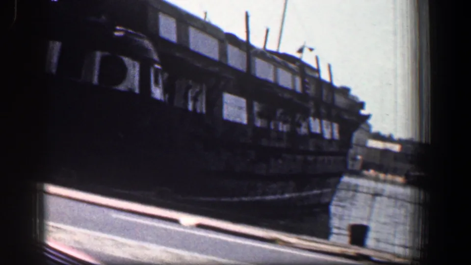 1961 Baltimore harbor view with a dark docked vessel, water, roadway edge, and film gate visible from a moving vehicle.