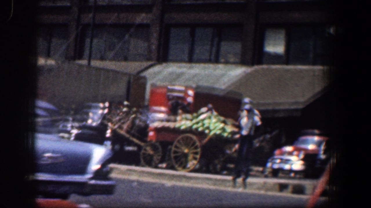 Horse-drawn produce cart on a city street with pedestrians and parked cars.