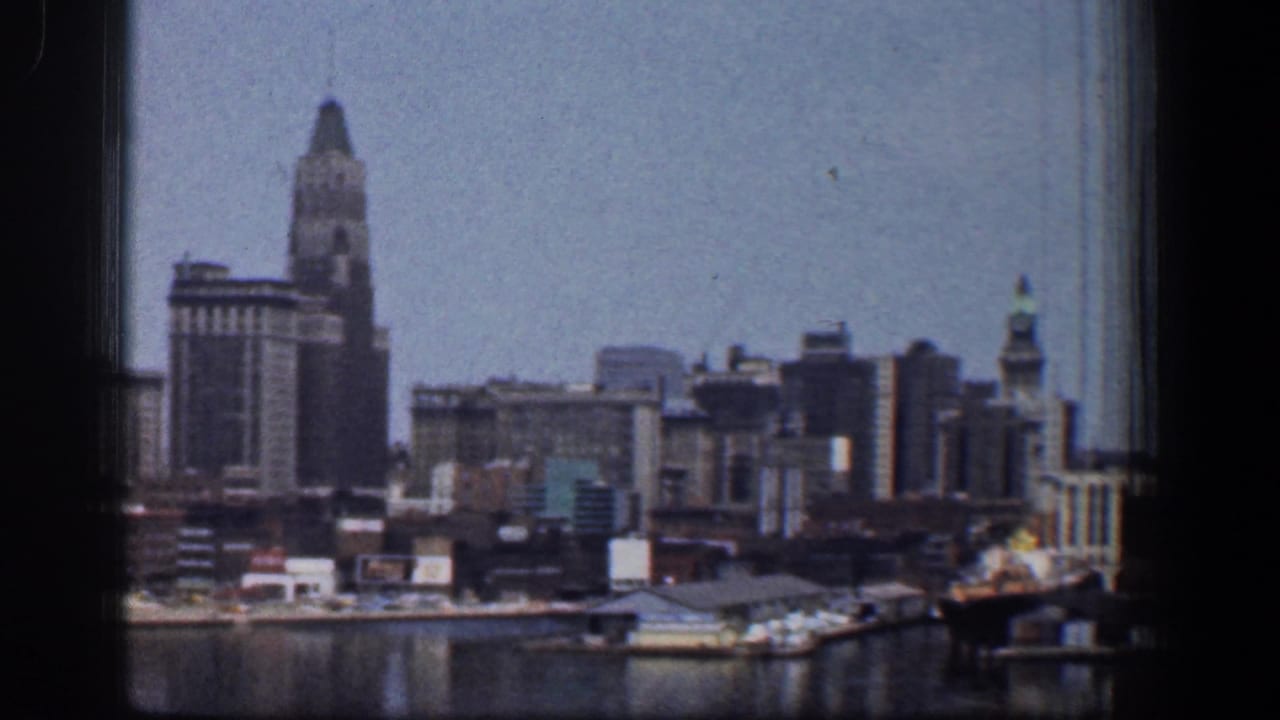 1961 Baltimore waterfront skyline on 8mm film, framed by a dark window edge with harbor water and dock structures in the foreground.