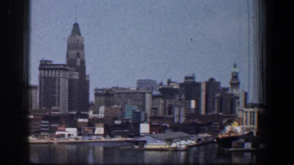 1961 Baltimore waterfront skyline on 8mm film, framed by a dark window edge with harbor water and dock structures in the foreground.