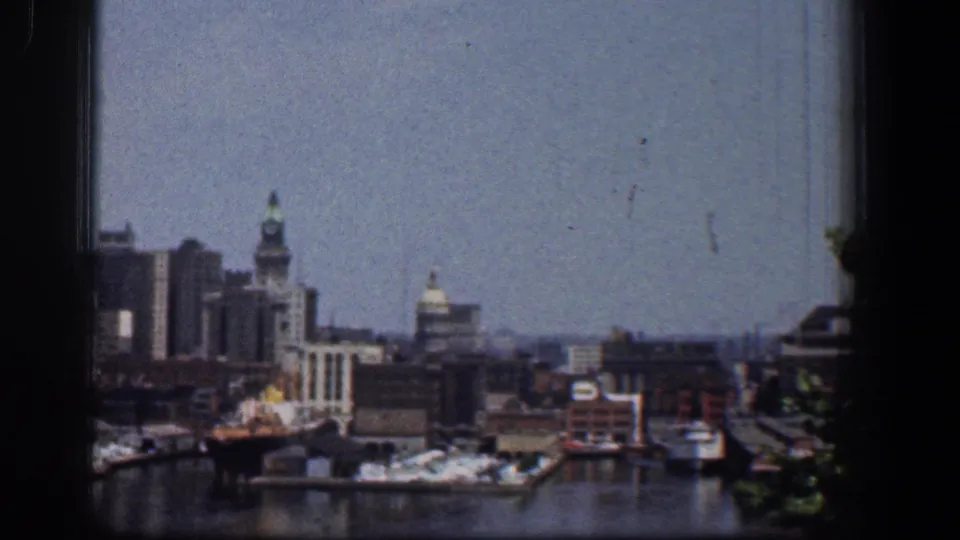 1961 Baltimore harbor basin with docked boats, waterfront buildings, and a distant city skyline in soft 8mm color.