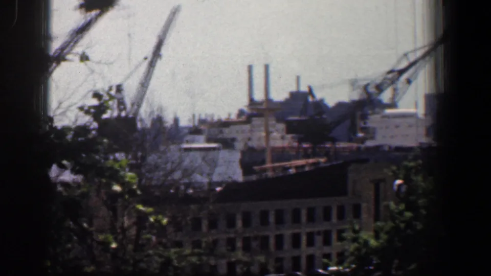 1961 Baltimore industrial waterfront view with cranes, factory stacks, low buildings, and foreground trees seen in soft home-movie focus.