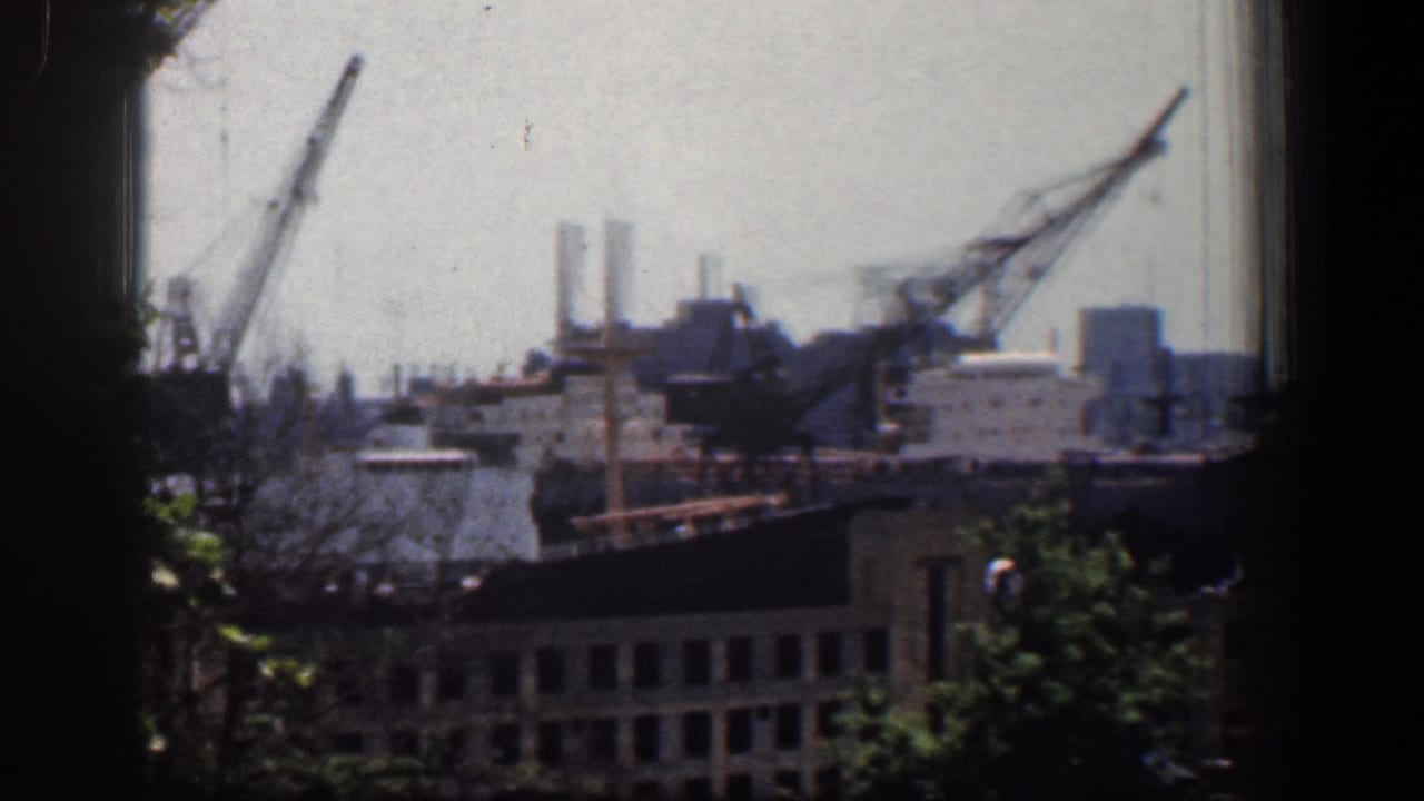 Industrial waterfront seen through a dark window frame, with cranes, buildings, trees, and rooftops.