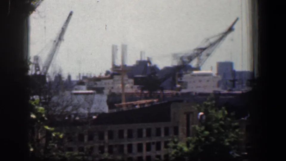 1961 Baltimore industrial waterfront viewed through a window frame, with cranes rising above low buildings and trees in the foreground.
