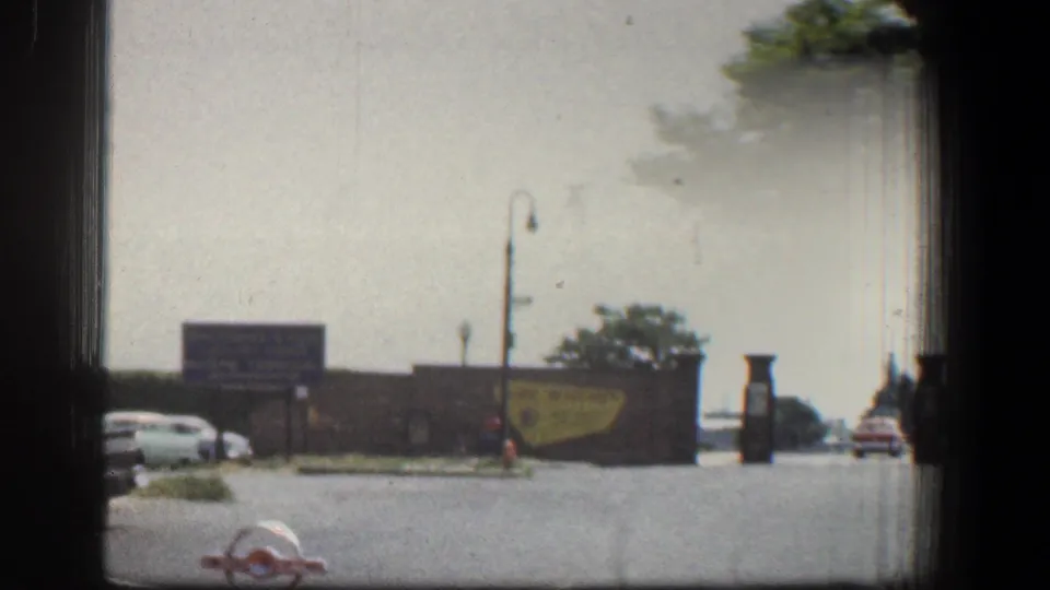 1961 Baltimore street-level waterfront entrance with period cars, signs, lamps, trees, and a glimpse of water beyond the wall.