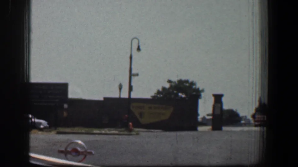 A 1961 Baltimore waterfront entrance area shows signs, lamps, trees, a parked or passing car, and small distant people near the street.