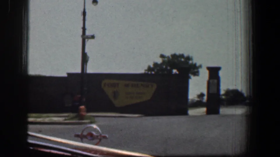 1961 Baltimore car-window view with a yellow Fort McHenry sign, street fixtures, and a roadside edge in soft 8mm color.
