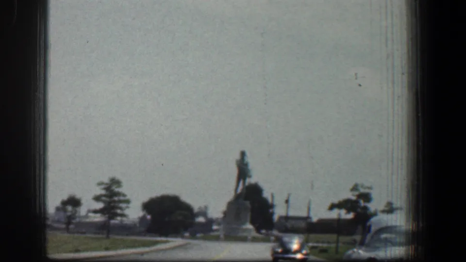 1961 Baltimore 8mm view of a distant statue near a roadway, with cars, trees, and lawn visible from a moving vehicle.