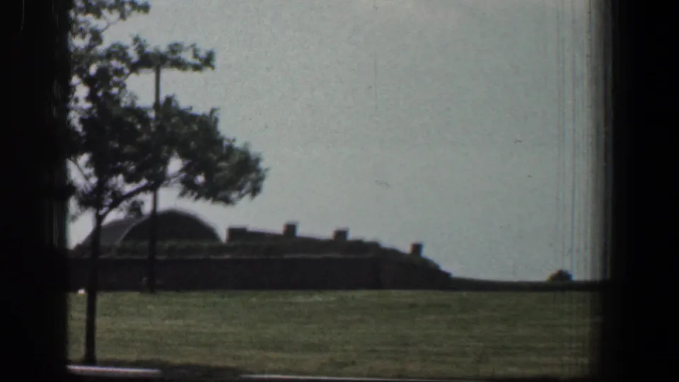 1961 Baltimore 8mm frame showing a grassy area, tree, low brown building, and dark window edge under a pale sky.