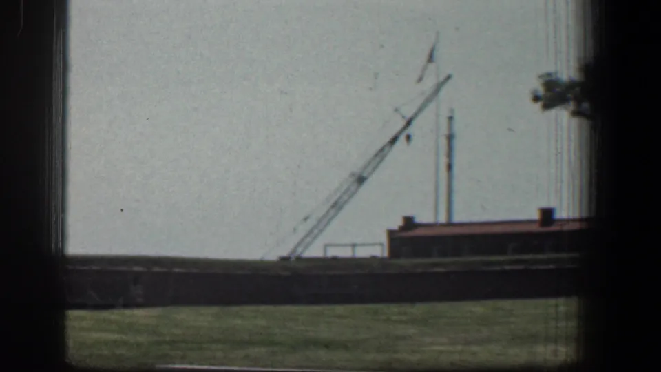 1961 Baltimore 8mm frame showing a crane near a low brown building, with grass in front and dark window framing.