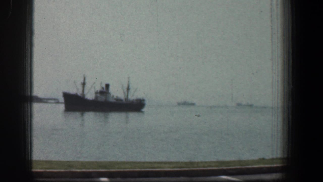 1961 Baltimore harbor view showing a large dark-hulled vessel on calm water, filmed through a window frame with distant boats on the horizon.