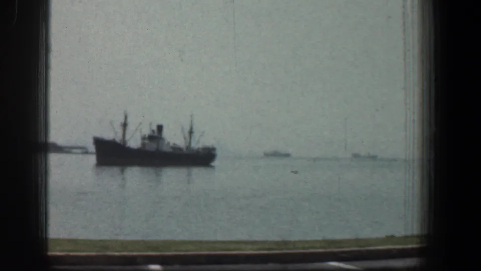 1961 Baltimore harbor view showing a large dark-hulled vessel on calm water, filmed through a window frame with distant boats on the horizon.
