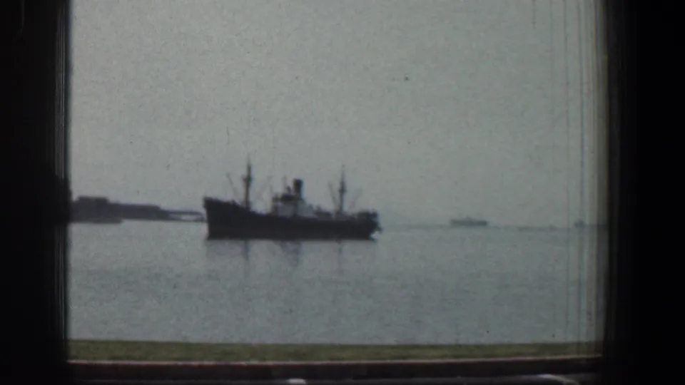 Large vessel on Baltimore harbor water in 1961, viewed through dark window framing with a low shoreline and small distant boats behind it.