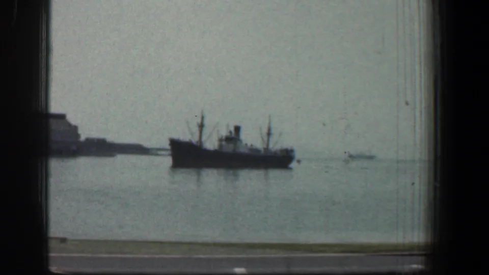 1961 Baltimore waterfront shot with a large vessel on calm harbor water, distant shoreline at left, and dark window edges around the frame.