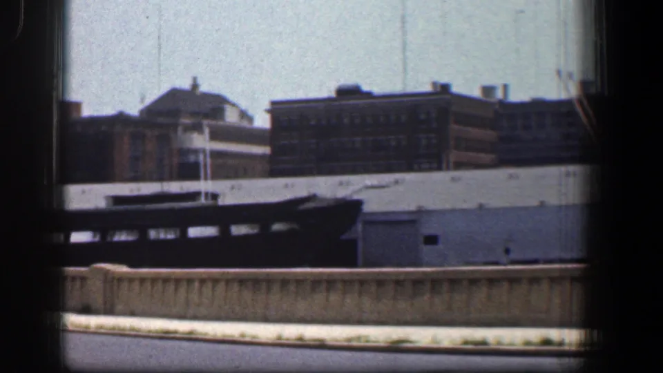 Baltimore industrial waterfront in 1961 with brick and warehouse-like buildings, a dark vessel in the foreground, and a low waterfront barrier seen through window framing.