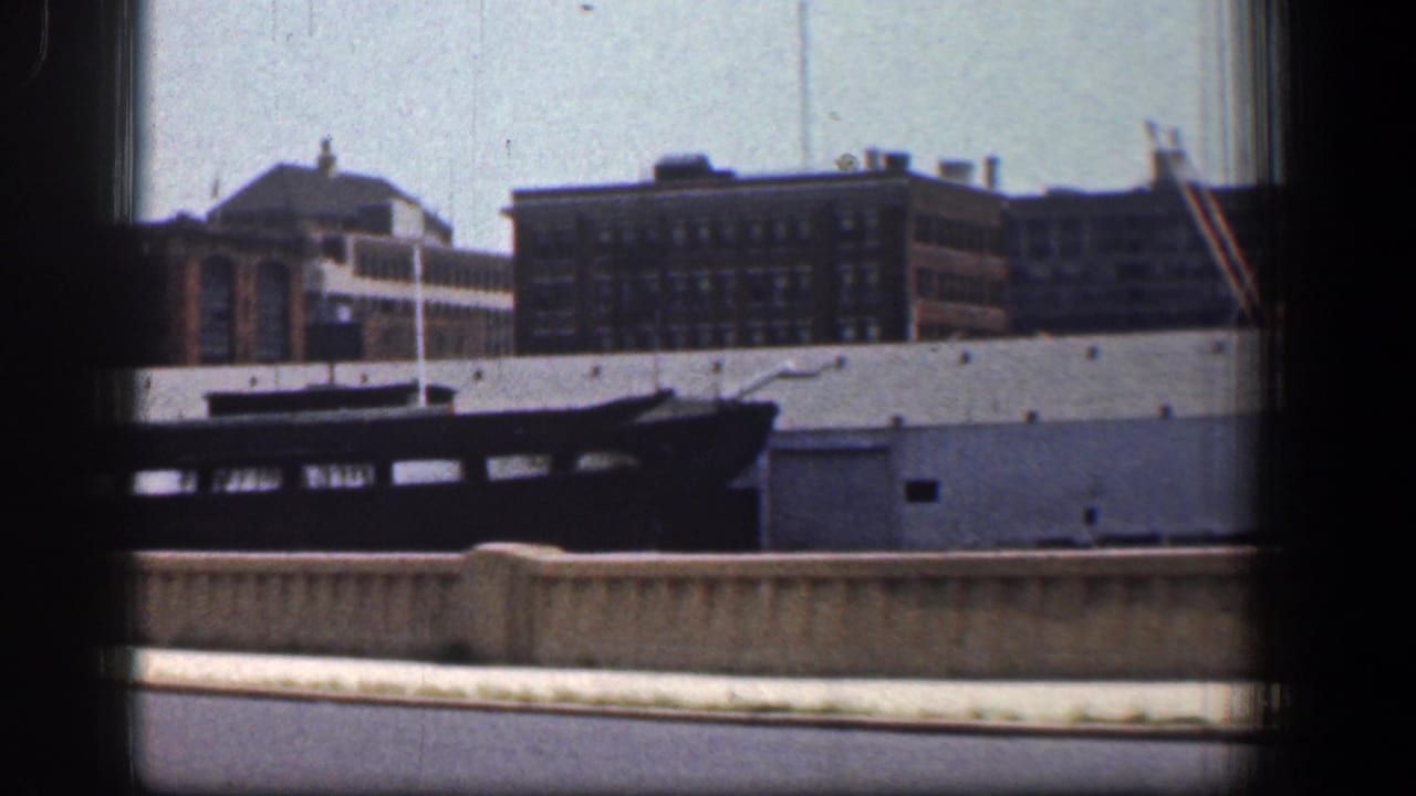 Dark boat along a waterfront with industrial buildings beyond a roadway barrier.