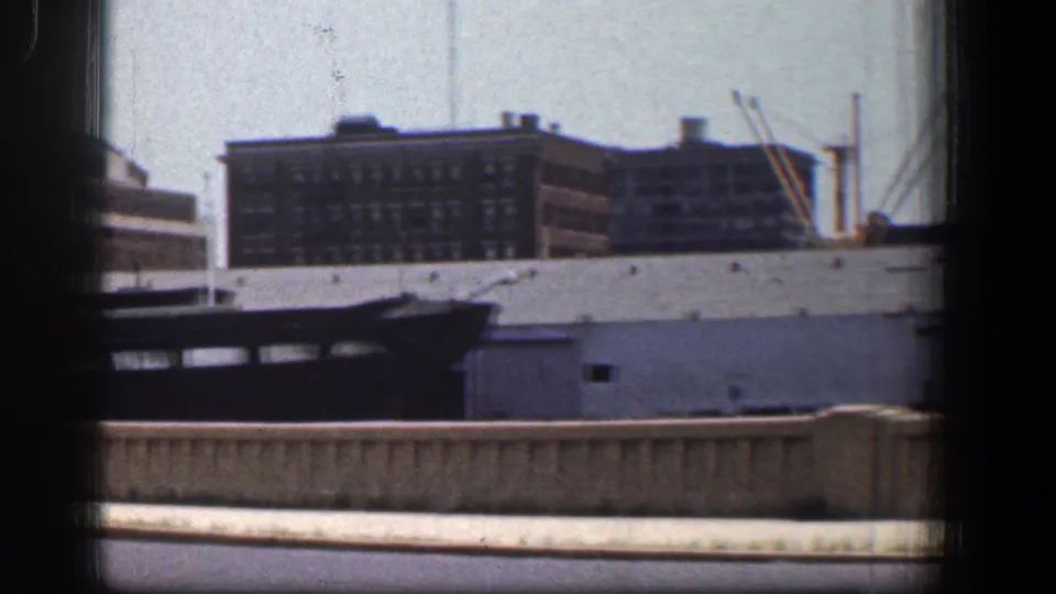 Moving 8mm view of a Baltimore waterfront with a dark boat, brick industrial buildings, and dock structures under a pale sky.