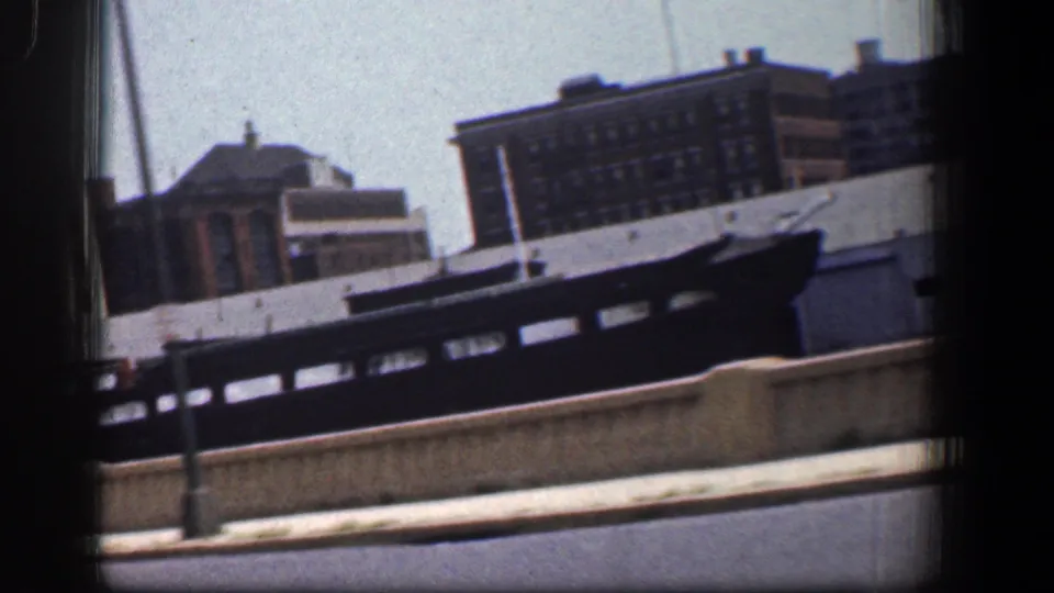 A canted 1961 Baltimore harbor frame shows the dark boat and waterfront buildings blurred slightly by vehicle motion.