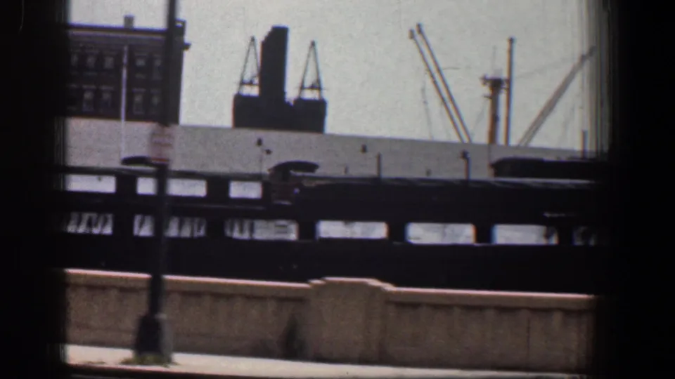 Baltimore industrial scene from a moving 8mm camera, showing cranes, dark rail or bridge structure, and distant masonry buildings.