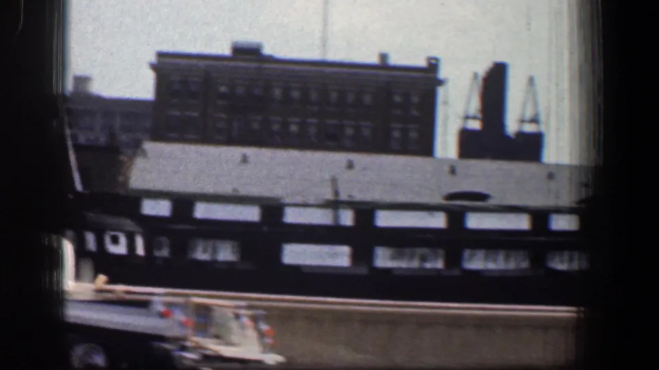 Moving 1961 Baltimore street view with a distant masonry building, cranes, elevated rail structure, and part of a car below.