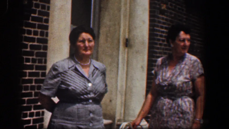 Two adult women in 1960s attire stand outside a brick building near a pale doorway, with faces visible.