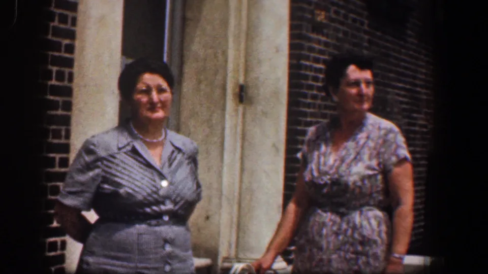 Two adult women pose outside a red brick building in Baltimore, Maryland, in 1961, one in a gray dress and pearls, the other in a floral dress.