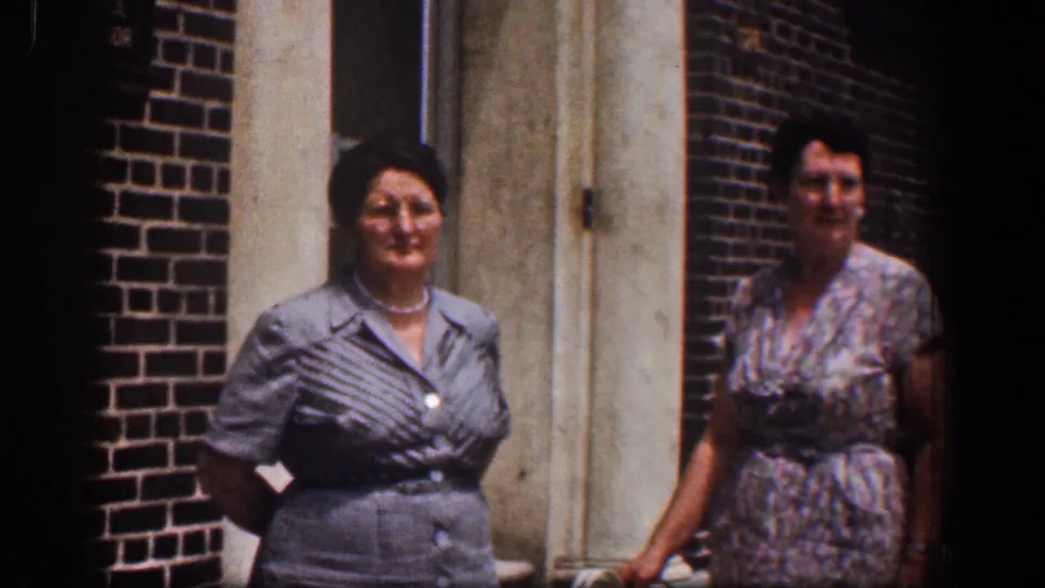 In a 1961 Baltimore exterior, two women stand near a brick building entrance, with the woman in gray facing the camera and the woman in floral print turned aside.