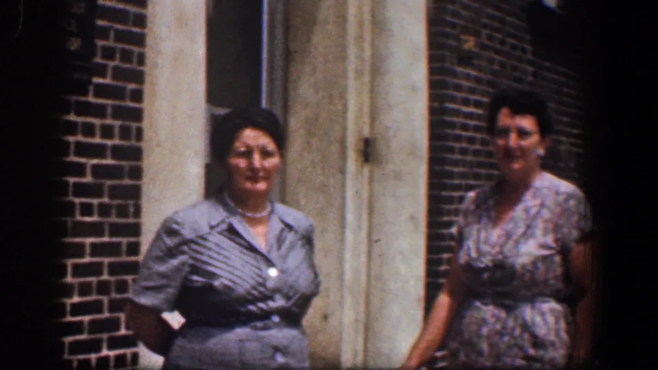 Two adult women stand outside a Baltimore brick building in 1961, the left woman centered in gray with pearls and the right woman partly turned in a floral dress.