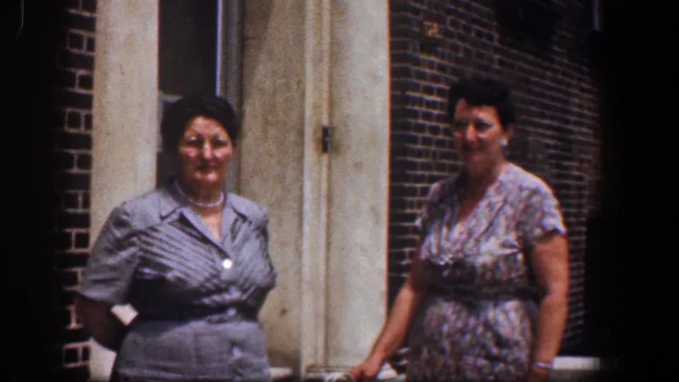 Near the end of the 1961 Baltimore clip, two women continue posing beside the brick exterior and doorway, with faces visible and clothing clearly shown.