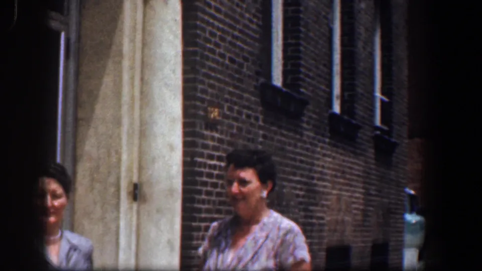 The women remain near the brick residential exterior, partly obscured by the doorway and film framing.