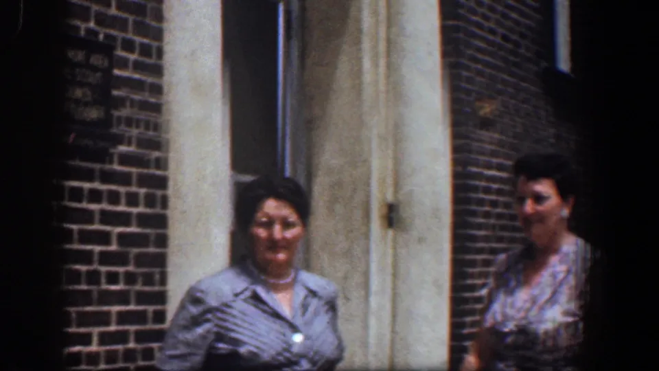 A closer home-movie view shows the women beside the brick wall and pale doorway, with soft focus and visible grain.