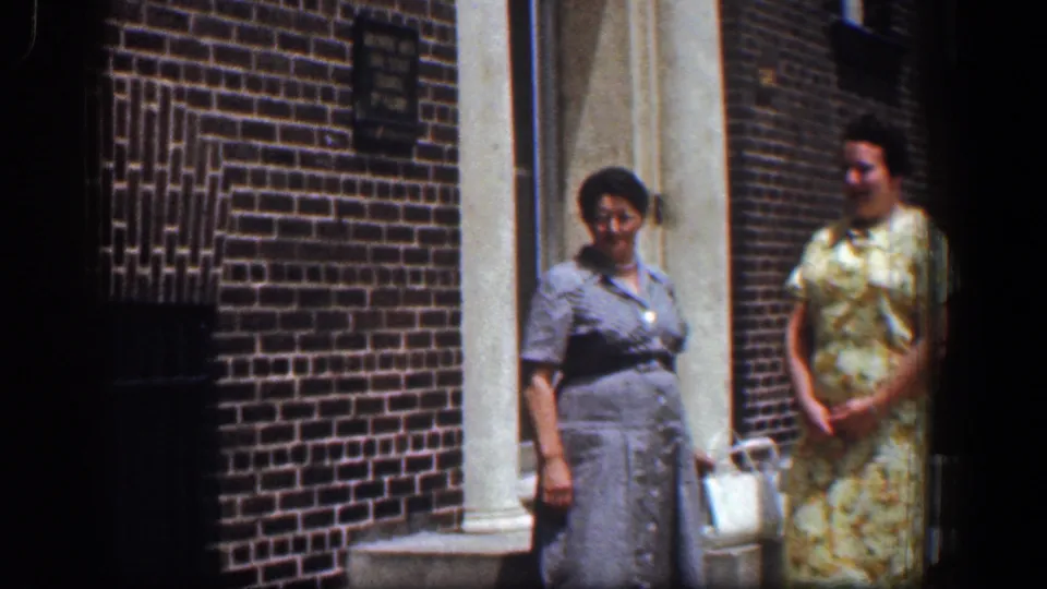 The women stand near a brick exterior and doorway, with handbags and dresses visible in the Baltimore home-movie scene.