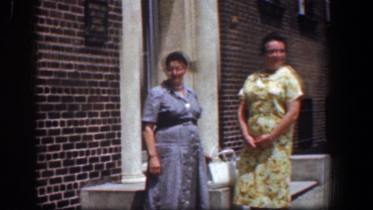 Two women stand by a red brick building entrance in bright sunlight.