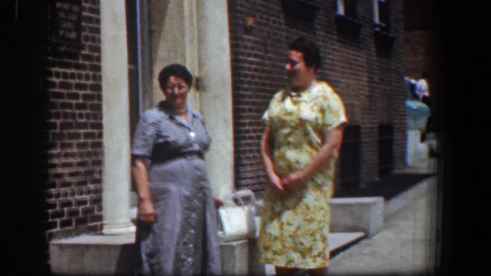 The women stand outside the Baltimore brick building, with the woman in gray near a white handbag and the woman in yellow turned slightly toward her.