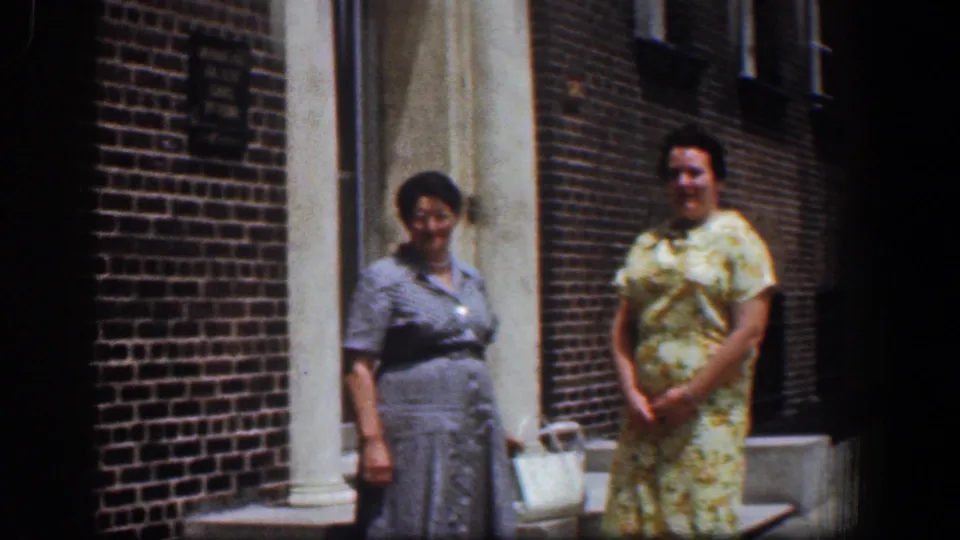 In the 1961 Baltimore home-movie frame, two women stand casually outside a red brick building with concrete steps and a pale entrance column.