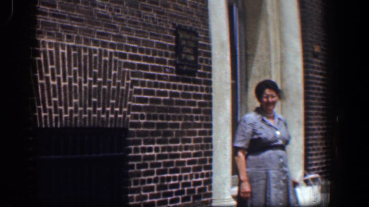In 1961 Baltimore footage, a woman in a gray dress stands near a white column outside a brick building entrance.