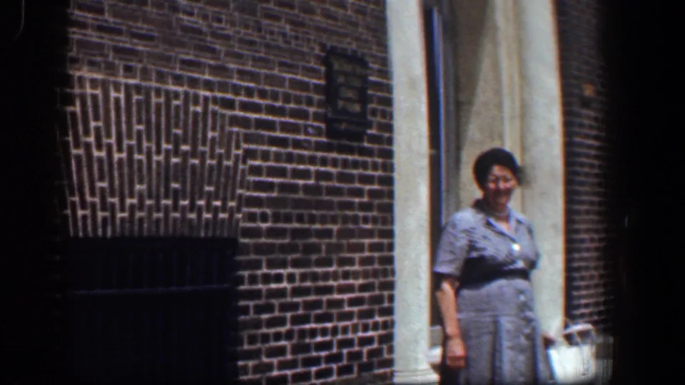 In 1961 Baltimore footage, a woman in a gray dress stands near a white column outside a brick building entrance.