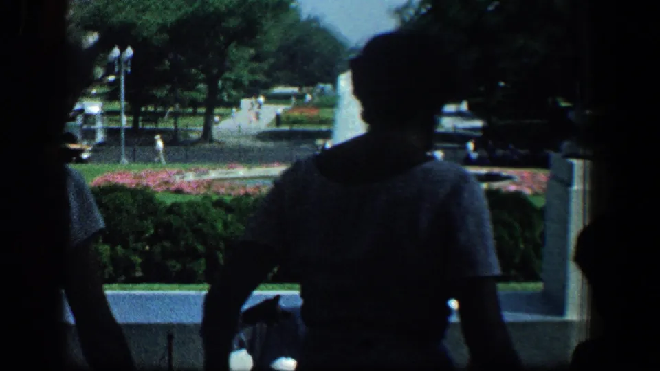 1961 Baltimore frame with a silhouetted person in a shaded doorway overlooking a sunlit park, trees, and flower beds.