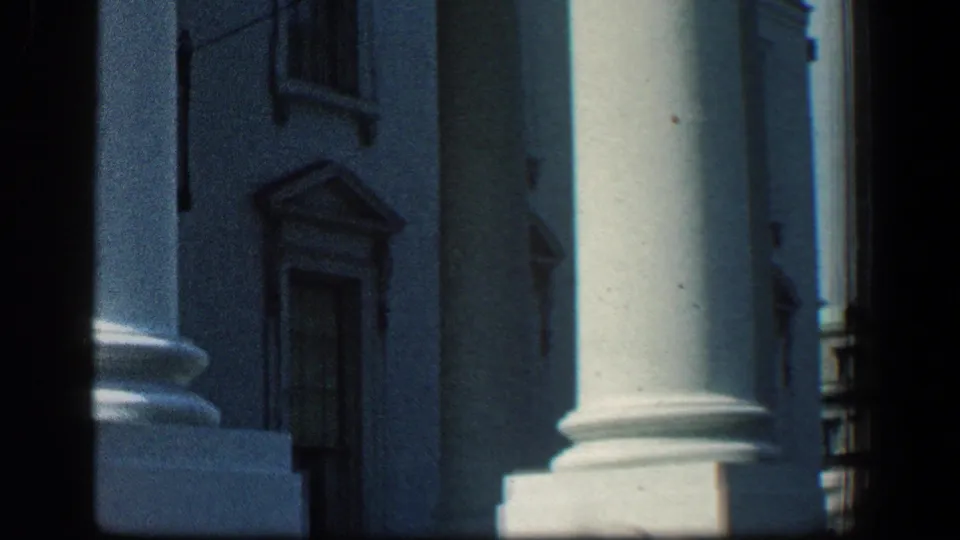 1961 Baltimore frame showing white classical columns, facade details, and strong shade along the building exterior.