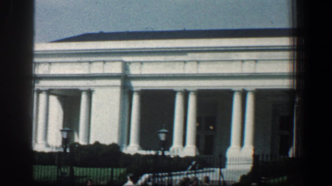 White colonnaded building seen beyond lawn, railings, and pedestrians.
